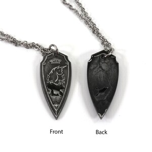 May include: A silver chain necklace with a pendant shaped like a shield. The front of the pendant features a detailed design of a unicorn with a crown above it. The back of the pendant is plain.