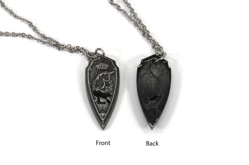 May include: A silver chain necklace with a pendant shaped like a shield. The front of the pendant features a detailed design of a unicorn with a crown above it. The back of the pendant is plain.