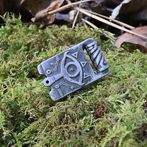 May include: A silver metal charm with a detailed design of an eye with triangles and a spiral pattern. The charm is on a bed of green moss.