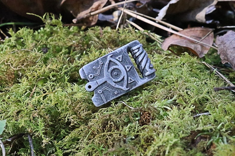 May include: A silver metal charm with a detailed design of an eye with triangles and a spiral pattern. The charm is on a bed of green moss.