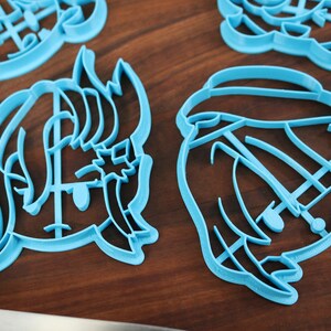 May include: Four blue plastic cookie cutters shaped like cartoon characters. The cutters are on a wooden surface.