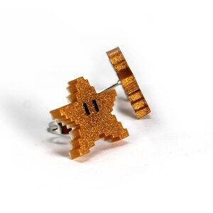 May include: A pair of gold-colored earrings featuring a pixelated star and a circular design. The star has a black face with two eyes.