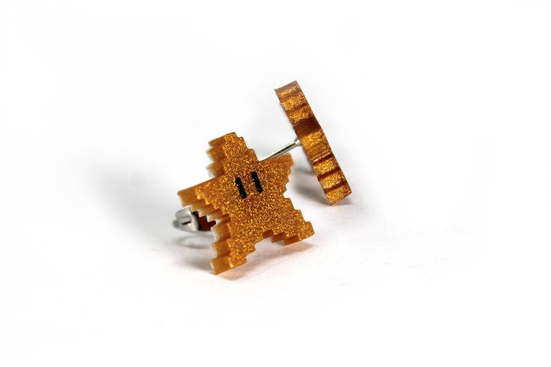 May include: A pair of gold-colored earrings featuring a pixelated star and a circular design. The star has a black face with two eyes.