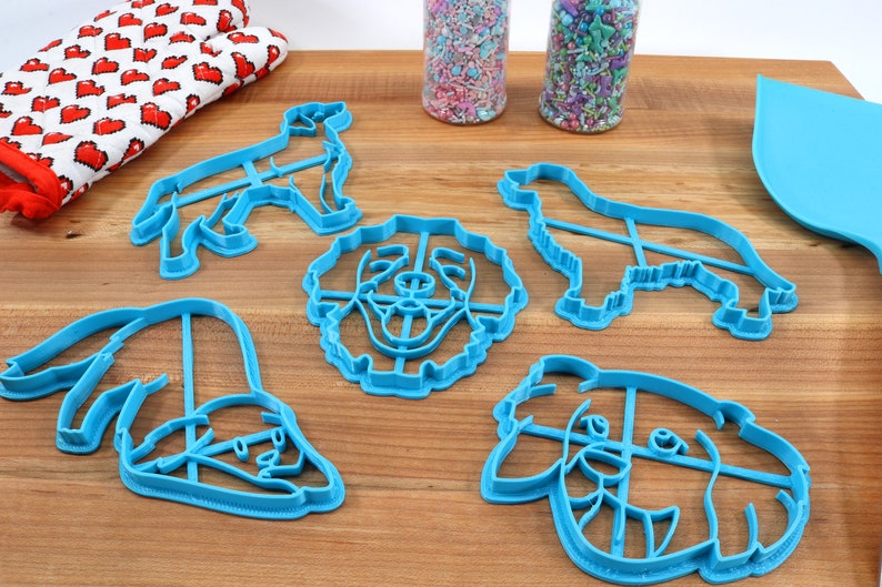 May include: A set of five blue plastic dog-shaped cookie cutters. The cutters are in the shape of a dog's head, a dog's body, and a dog's paw.