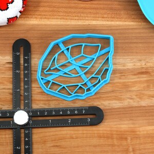 May include: A blue plastic leaf-shaped cookie cutter. The cutter has a detailed design with a raised edge.