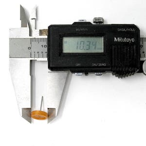 May include: A silver caliper with a black digital display reading 10.34 mm. The caliper is measuring a small, round, orange object.