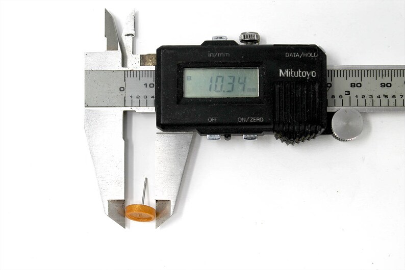 May include: A silver caliper with a black digital display reading 10.34 mm. The caliper is measuring a small, round, orange object.