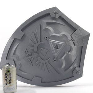 May include: A gray plastic replica of the Hylian Shield from the Legend of Zelda video game series. The shield features a raised design of the Triforce and the Hylian crest. The shield is approximately 12 inches in diameter.