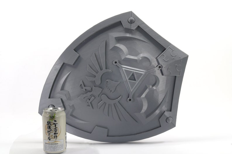 May include: A gray plastic replica of the Hylian Shield from the Legend of Zelda video game series. The shield features a raised design of the Triforce and the Hylian crest. The shield is approximately 12 inches in diameter.