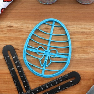 May include: A blue plastic cookie cutter shaped like an Easter egg with a bow on it. The cutter is on a wooden surface.