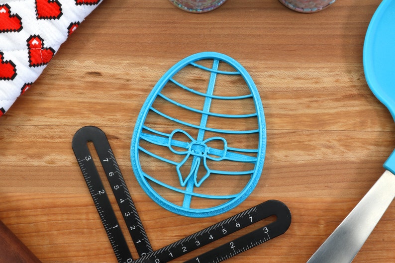 May include: A blue plastic cookie cutter shaped like an Easter egg with a bow on it. The cutter is on a wooden surface.