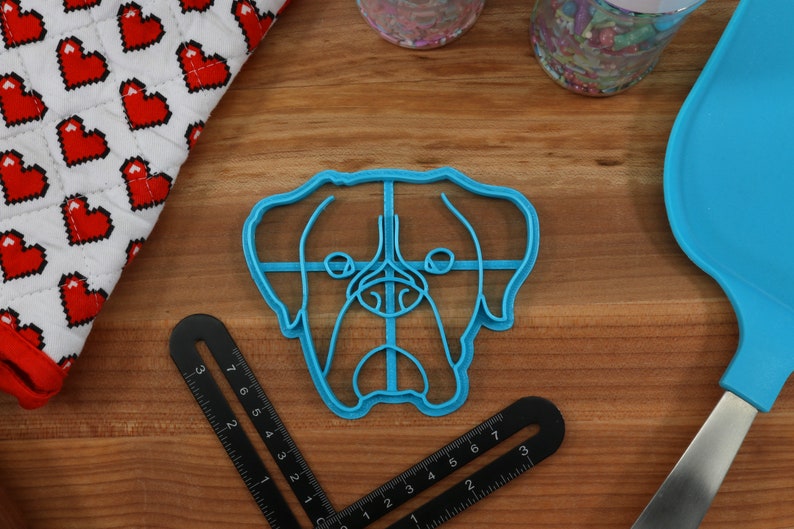 May include: A blue plastic cookie cutter shaped like a dog's head. The cutter has a detailed outline of the dog's face, including the eyes, nose, and mouth.