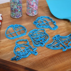 May include: A set of five blue plastic cookie cutters shaped like cartoon characters. The cutters are on a wooden surface with a rolling pin, a spatula, and a red oven mitt.