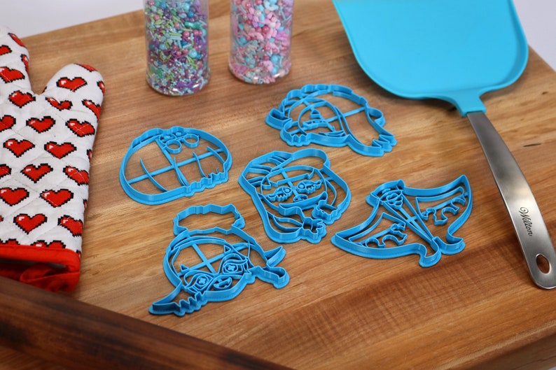 May include: A set of five blue plastic cookie cutters shaped like cartoon characters. The cutters are on a wooden surface with a rolling pin, a spatula, and a red oven mitt.