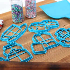 May include: A set of blue plastic cookie cutters shaped like a teapot, a house, a tree, a milk jug, and a box. The cutters are on a wooden cutting board with a spatula and two jars of sprinkles.