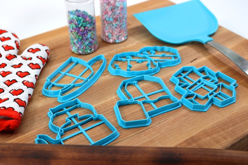 May include: A set of blue plastic cookie cutters shaped like a teapot, a house, a tree, a milk jug, and a box. The cutters are on a wooden cutting board with a spatula and two jars of sprinkles.