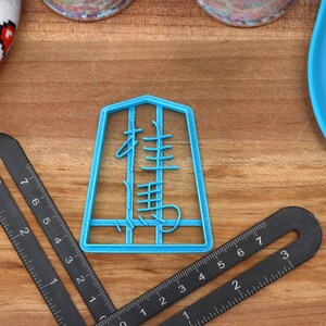 May include: A light blue cookie cutter in the shape of a Chinese character, with a black ruler and a heart-patterned oven mitt on a wooden surface. The ruler is marked with inches.