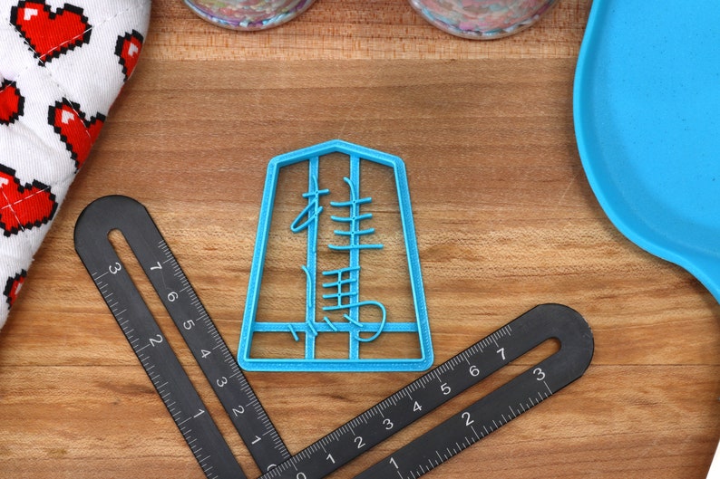 May include: A light blue cookie cutter in the shape of a Chinese character, with a black ruler and a heart-patterned oven mitt on a wooden surface. The ruler is marked with inches.