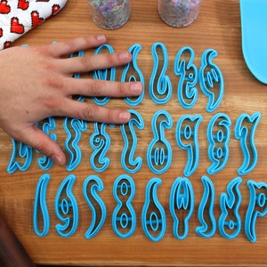 May include: A set of blue plastic cookie cutters shaped like different letters of the alphabet. The cutters are arranged in three rows on a wooden surface.