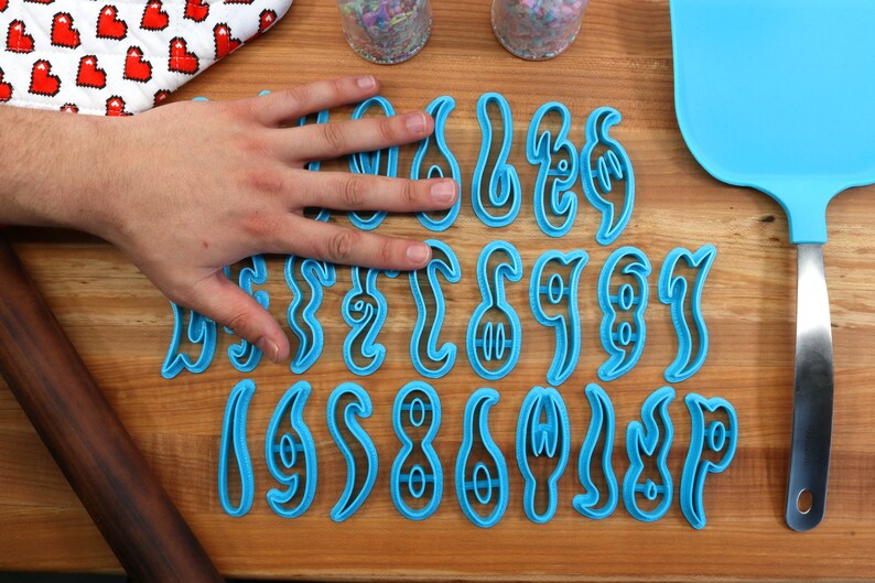 May include: A set of blue plastic cookie cutters shaped like different letters of the alphabet. The cutters are arranged in three rows on a wooden surface.