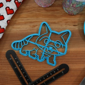 Puede incluir: Un cortador de galletas de pl&aacute;stico azul con forma de mapache de dibujos animados. El mapache est&aacute; sonriendo y tiene una cola rayada.
