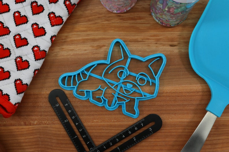 Puede incluir: Un cortador de galletas de pl&aacute;stico azul con forma de mapache de dibujos animados. El mapache est&aacute; sonriendo y tiene una cola rayada.