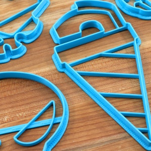 May include: A set of blue plastic cookie cutters in various shapes, including a skull, a cone, a circle, and a crescent moon. The cutters are on a wooden surface.