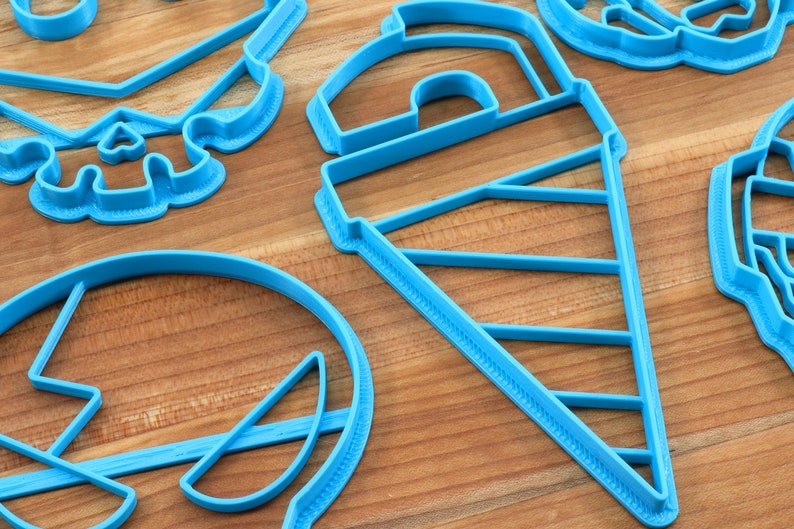 May include: A set of blue plastic cookie cutters in various shapes, including a skull, a cone, a circle, and a crescent moon. The cutters are on a wooden surface.