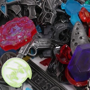 May include: A collection of colorful plastic and metal charms, including a red and purple charm with a starburst design, a silver gun charm, a blue and green charm with a geometric design, and a purple gem charm.