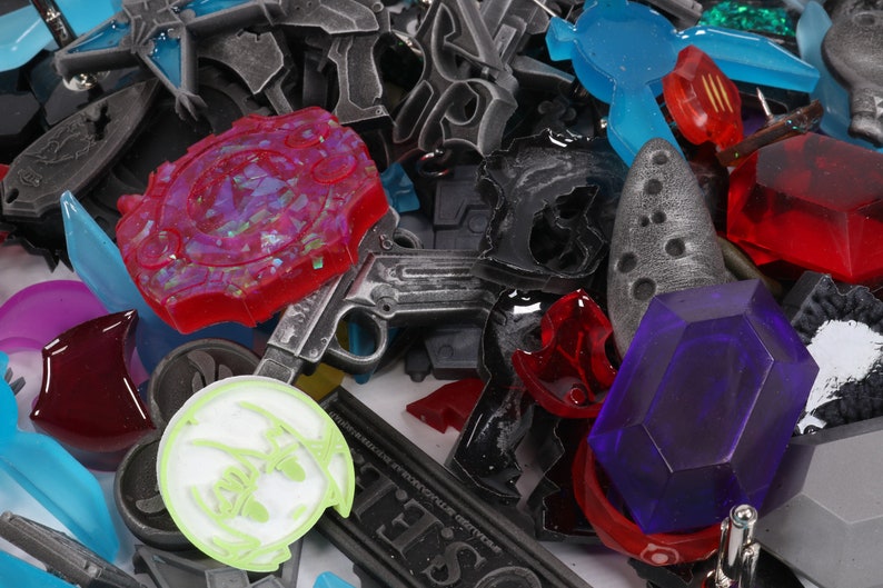 May include: A collection of colorful plastic and metal charms, including a red and purple charm with a starburst design, a silver gun charm, a blue and green charm with a geometric design, and a purple gem charm.