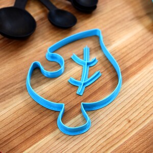May include: A blue plastic cookie cutter shaped like a leaf with a smaller leaf design inside. The cutter is on a wooden surface.