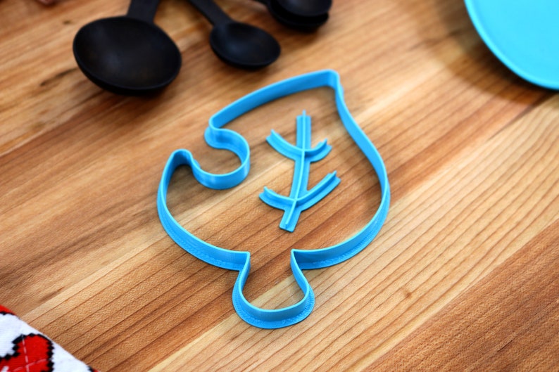 May include: A blue plastic cookie cutter shaped like a leaf with a smaller leaf design inside. The cutter is on a wooden surface.