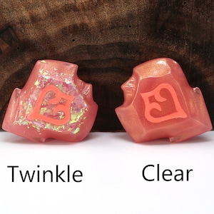 May include: Two pink resin dice with a number 5 on each face. The die on the left has a glittery finish, while the die on the right has a clear finish. The dice are labelled "Twinkle" and "Clear".