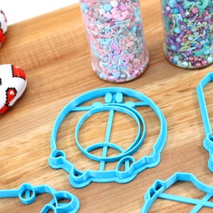 May include: A set of blue plastic cookie cutters shaped like a donut, a swing set, and a house. The cutters are on a wooden surface.