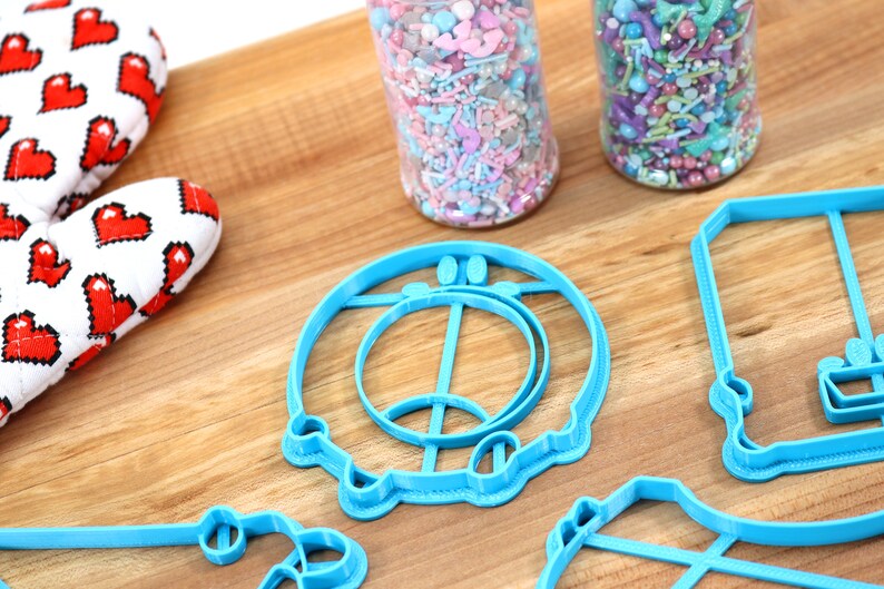 May include: A set of blue plastic cookie cutters shaped like a donut, a swing set, and a house. The cutters are on a wooden surface.
