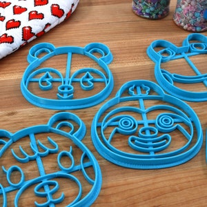 May include: Five blue plastic cookie cutters shaped like animal faces. The cutters are on a wooden surface. The cutters are for making cookies in the shape of a panda, a frog, a sloth, a bear, and a raccoon.