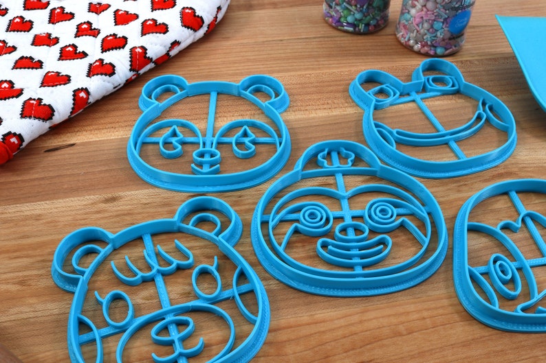 May include: Five blue plastic cookie cutters shaped like animal faces. The cutters are on a wooden surface. The cutters are for making cookies in the shape of a panda, a frog, a sloth, a bear, and a raccoon.