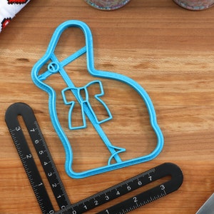May include: A blue plastic cookie cutter shaped like a bunny rabbit with a bow. The cutter is on a wooden surface.
