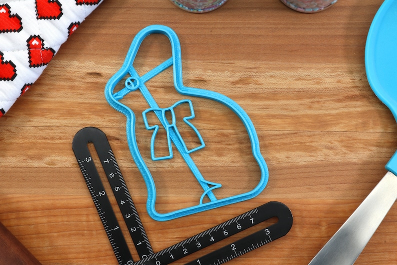 May include: A blue plastic cookie cutter shaped like a bunny rabbit with a bow. The cutter is on a wooden surface.
