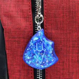 May include: A blue resin charm with a white design of a stylized hand with three fingers. The charm is attached to a silver zipper pull on a red fabric bag.