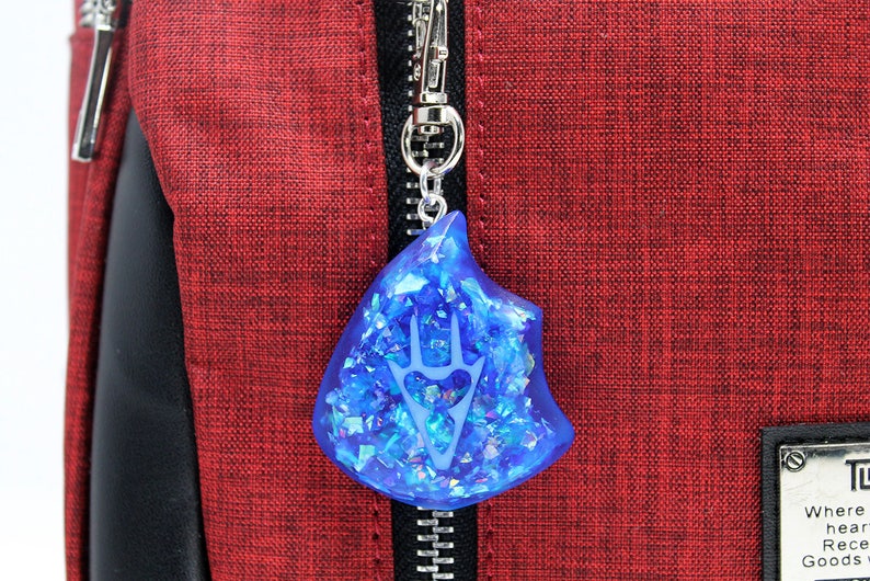 May include: A blue resin charm with a white design of a stylized hand with three fingers. The charm is attached to a silver zipper pull on a red fabric bag.