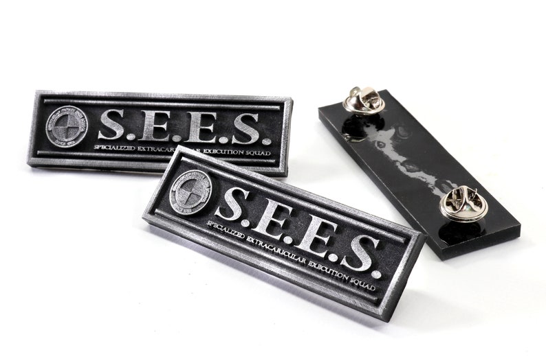 May include: Two silver metal pins with the text "S.E.E.S. SPECIALIZED EXTRACURRICULAR EXECUTION SQUAD" on a black background. The pins are on a white surface.