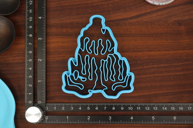 Puede incluir: Un cortador de galletas de pl&aacute;stico azul con forma de &aacute;rbol estilizado con un dise&ntilde;o ondulado y org&aacute;nico. El cortador est&aacute; sobre una superficie de madera.