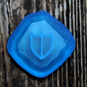 Puede incluir: Una pieza de juego de vidrio azul con un dise&ntilde;o de escudo. El escudo es blanco y tiene tres puntos en la parte superior.