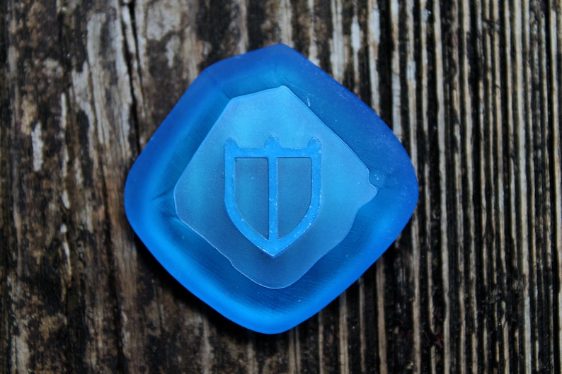 Puede incluir: Una pieza de juego de vidrio azul con un dise&ntilde;o de escudo. El escudo es blanco y tiene tres puntos en la parte superior.