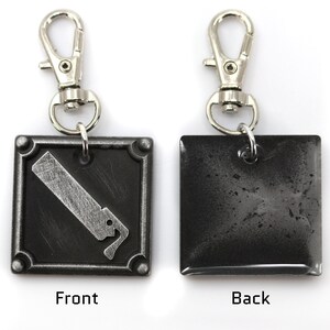 May include: A silver keychain with a black square charm. The front of the charm has a silver graphic of a saw blade. The back of the charm is plain.
