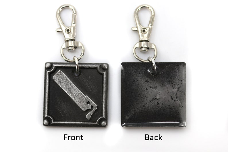 May include: A silver keychain with a black square charm. The front of the charm has a silver graphic of a saw blade. The back of the charm is plain.