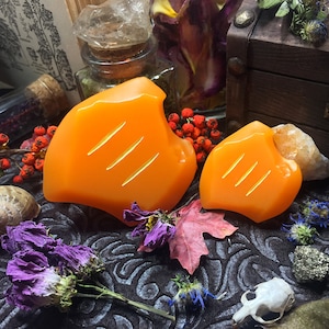 May include: Two orange, hand-carved soap bars with a textured surface. The soap bars are shaped like a leaf and have three lines carved into the surface. The soap bars are sitting on a dark brown surface with dried flowers and other decorative items.