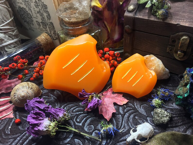 May include: Two orange, hand-carved soap bars with a textured surface. The soap bars are shaped like a leaf and have three lines carved into the surface. The soap bars are sitting on a dark brown surface with dried flowers and other decorative items.