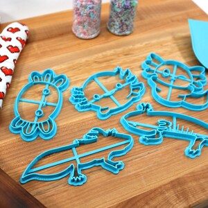 May include: Five blue plastic cookie cutters shaped like axolotls, a turtle, and a hedgehog on a wooden cutting board. There are two jars of sprinkles in the background.